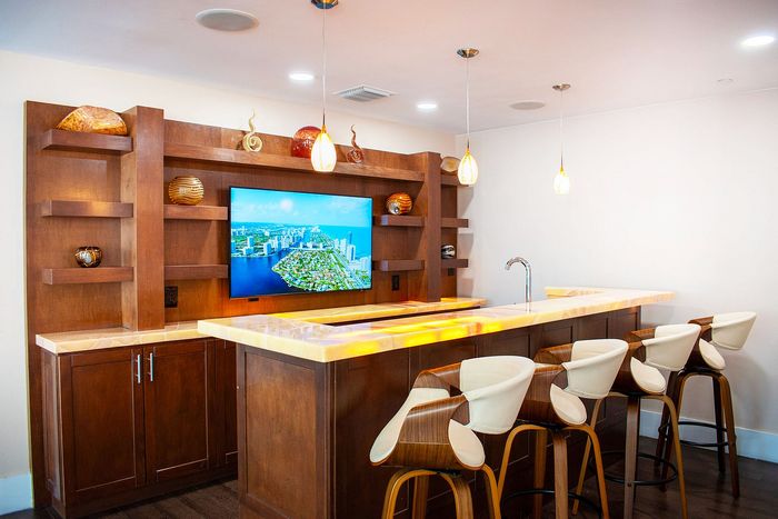 Modern home bar with elegant wooden cabinetry, illuminated countertops, stylish bar stools, and a wall-mounted TV displaying a vibrant cityscape.