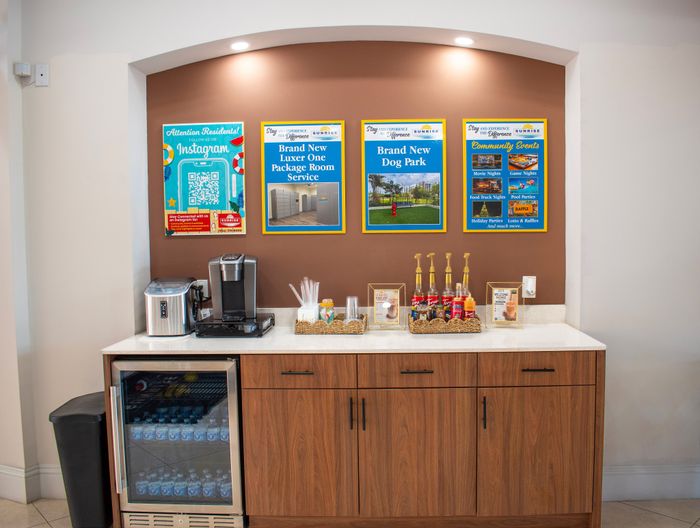 Inviting apartment amenities area featuring a coffee station with syrup selections and a mini fridge, complemented by colorful community event posters on a stylish accent wall.