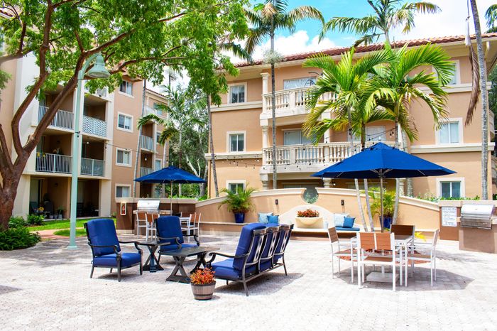 Charming apartment complex courtyard featuring elegant outdoor seating, lush palm trees, and vibrant blue umbrellas on a sunny day.
