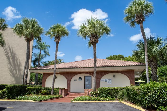Charming community entrance with classic red brick architecture and lush palm trees under a bright blue sky.