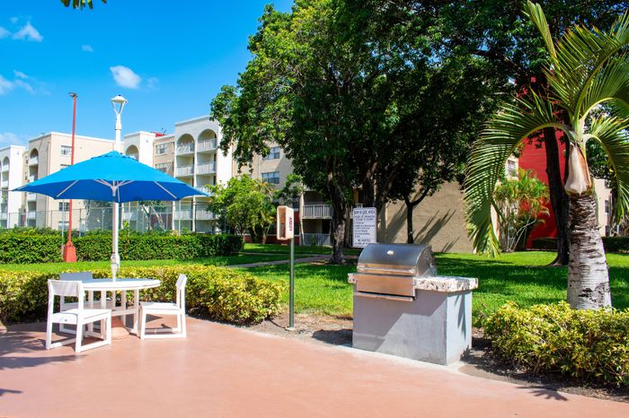 Lush outdoor area with modern barbecue setup, shaded seating under blue umbrella, surrounded by vibrant greenery and apartment buildings.