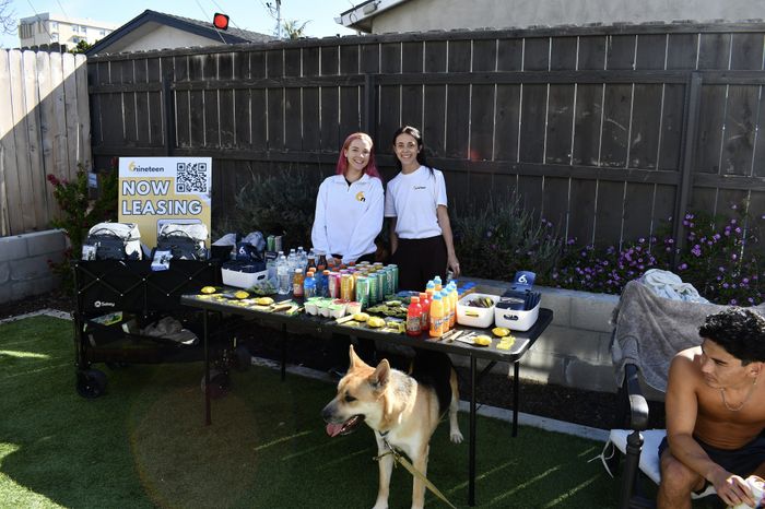 Two women stand behind a leasing table full of drinks, snacks, and giveaways, smiling at the camera. A German Shepherd dog stands in front of them on a grassy backyard lawn.