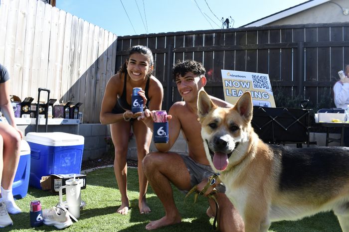 A young man and woman kneel on the grass next to a smiling dog, holding branded drink koozies. They pose proudly in front of a leasing booth under a sunny sky with