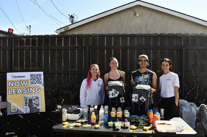 Four young adults, two holding branded backpacks, pose and smile behind a leasing booth table stocked with drinks, snacks, and giveaway items. A