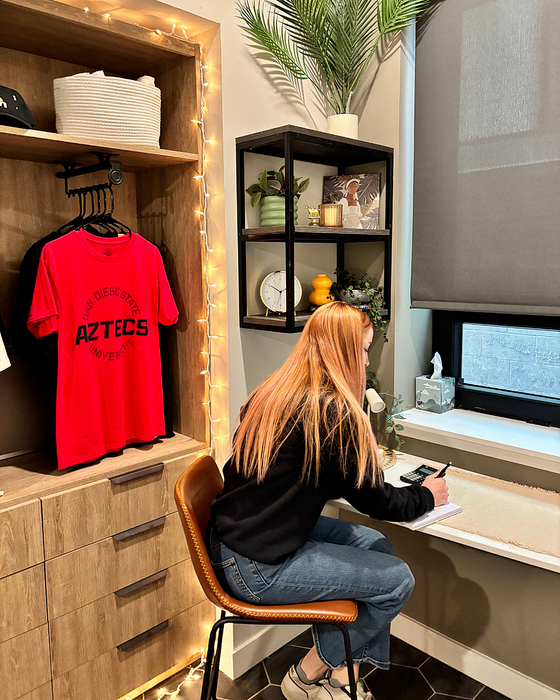 A red-haired woman sits at a small desk writing in a notebook, facing a window. The room features wooden furniture, a red San Diego State University shirt hanging in a closet, and decorative string lights. Shelves with plants, a clock, and a framed photo are mounted on the wall above her.