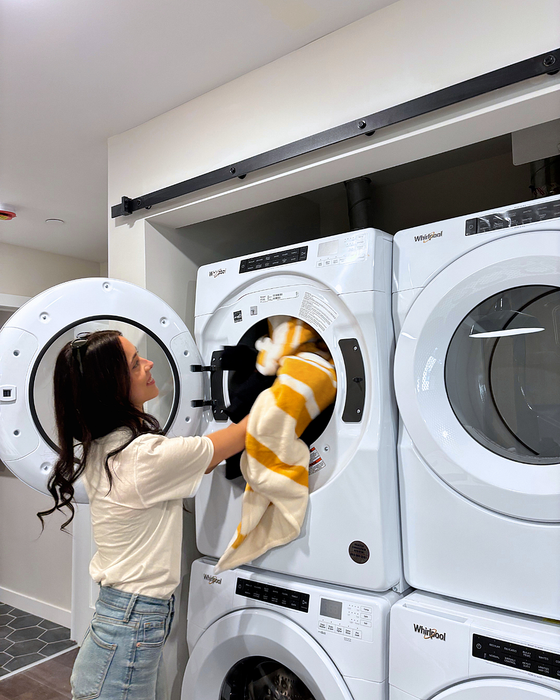 A woman with long dark hair loads a yellow and white striped blanket into a front-loading Whirlpool dryer in a communal laundry room. She&rsquo;s wearing a cream top and jeans, and the room has a clean, modern design with white machines and hexagon tile flooring.