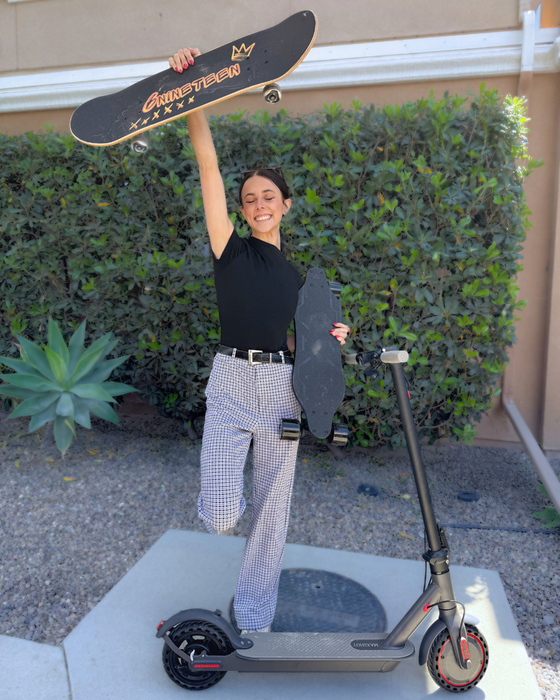A smiling woman stands on one foot next to an electric scooter, holding a skateboard in one hand and raising another skateboard over her head. She wears a black top and checkered pants, and the background features leafy green bushes.