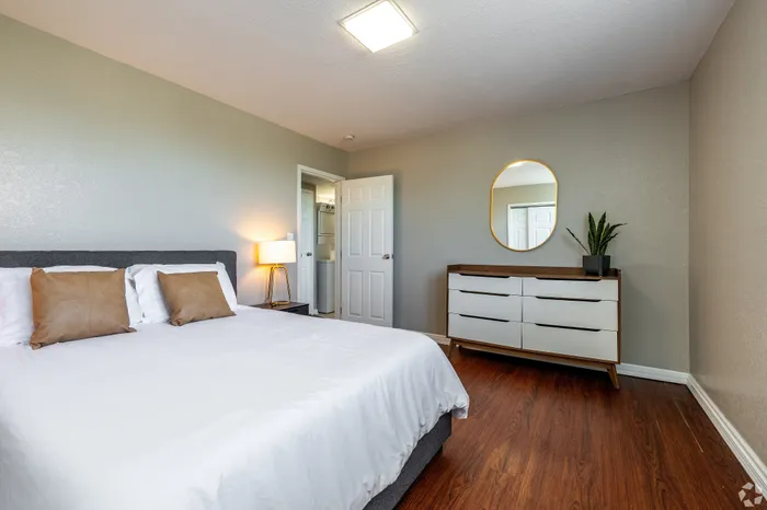 Bright apartment bedroom featuring a neatly made bed, wood-style flooring, a modern dresser with mirror, and an open doorway to an in-unit washer and dryer, offering a comfortable and functional living space.