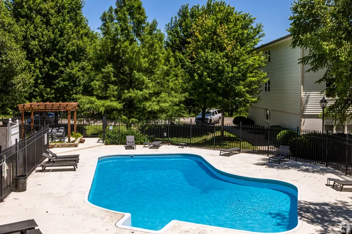 Resort-style outdoor swimming pool with lounge chairs, a shaded pergola, and secure fencing, surrounded by mature trees within a peaceful apartment community setting.