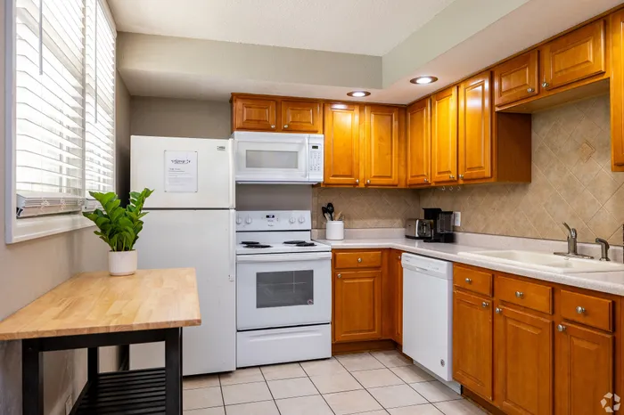 Apartment kitchen with warm wood cabinetry, white appliances, tiled backsplash, dishwasher, and a small breakfast table by the window, offering a functional and inviting space for everyday cooking and dining.