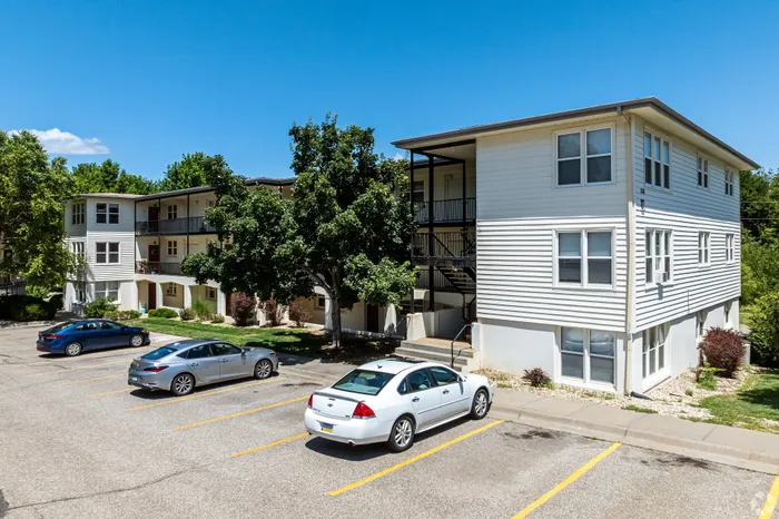 Exterior view of a garden-style apartment community with light-colored buildings, private balconies, mature trees, and on-site parking, offering a quiet residential setting with convenient access for residents.