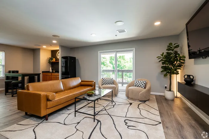 Modern apartment community lounge featuring a leather sofa, accent chairs, wall-mounted TV, and sliding doors to a balcony, creating a comfortable shared space for residents to relax and socialize.