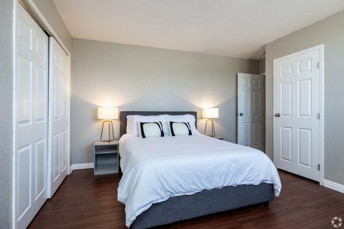 Bright apartment bedroom with a neatly made bed, wood-style flooring, bedside lamps, and double closets, offering a clean and comfortable space with ample storage.