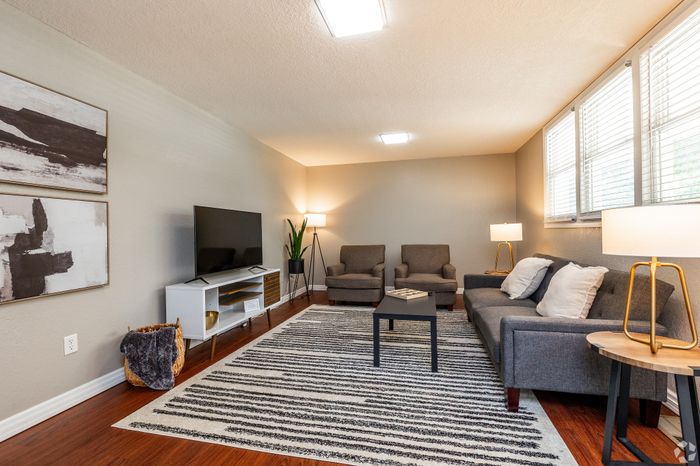 Spacious apartment living room with a sofa, accent chairs, wall-mounted TV, wood-style flooring, and large windows, creating a bright and inviting space for everyday living and entertaining.