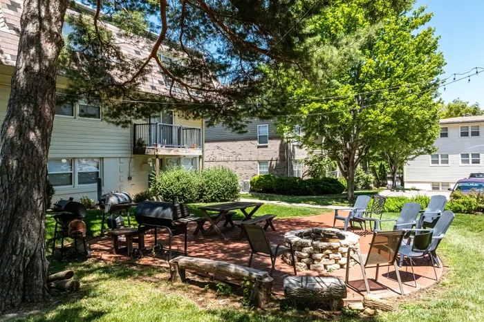 Outdoor apartment community courtyard with a fire pit, picnic tables, grilling stations, and lounge seating beneath mature trees, creating a welcoming space for residents to gather and relax outdoors.