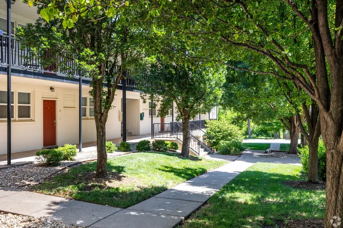 Tree-lined walkway through a landscaped apartment community with ground-level entrances, shaded sidewalks, and well-maintained green space, creating a quiet and welcoming residential atmosphere.