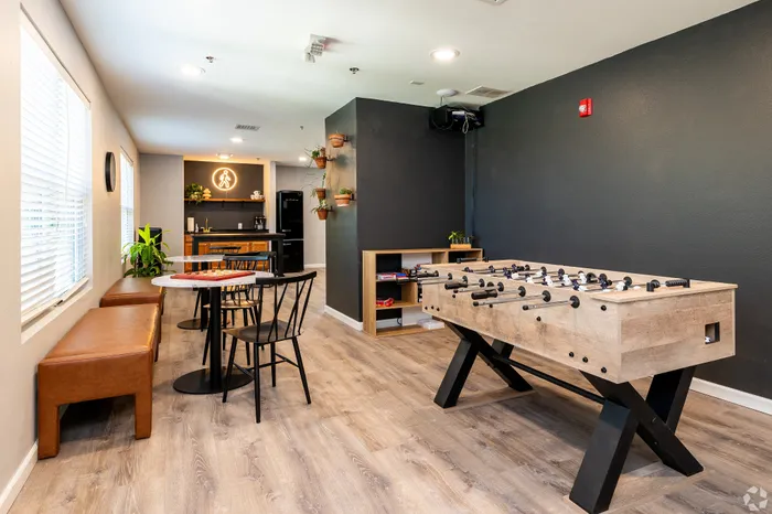 Apartment community game lounge with a foosball table, caf&eacute;-style seating, wall-mounted plants, and a kitchenette, offering a modern indoor space for residents to gather, play, and relax.