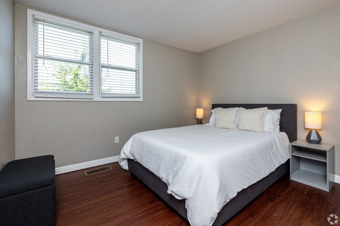 Bright apartment bedroom with a neatly made bed, wood-style flooring, bedside lamps, and a window with blinds, offering a clean and comfortable retreat with natural light.