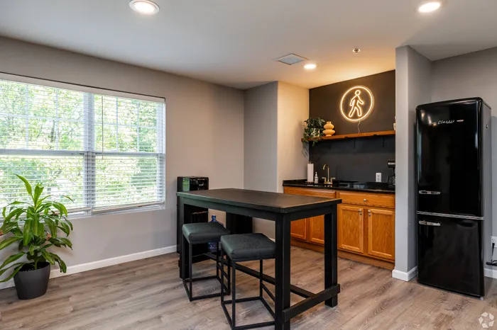 Apartment community kitchenette and gathering space with caf&eacute;-style table seating, wood cabinetry, black refrigerator, and modern lighting, offering a convenient spot for residents to grab refreshments and connect.