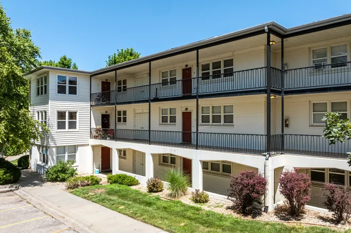 Exterior view of a garden-style apartment building with covered balconies, multiple entry doors, and landscaped greenery, offering a well-maintained community setting with outdoor access for residents.