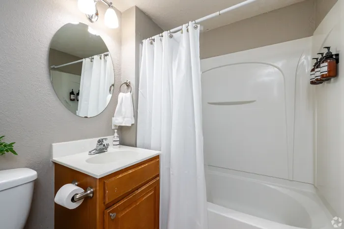 Apartment bathroom with a vanity sink, round mirror, tub-and-shower combination with curtain, and wood cabinetry, offering a clean and functional space designed for everyday comfort.