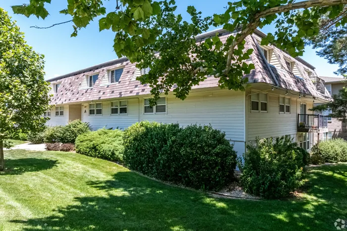 Exterior view of a garden-style apartment building with light siding, upper-level windows, and manicured landscaping, surrounded by mature trees and green lawn for a quiet residential feel.