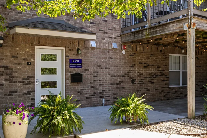 Exterior view of the Prime Place leasing office with a brick fa&ccedil;ade, modern entry door, shaded patio, and landscaped planters, creating a welcoming first impression for prospective residents.