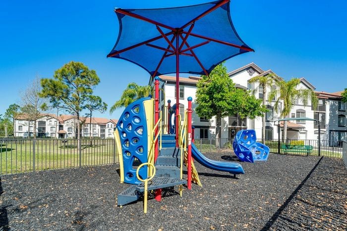 Vibrant playground with modern play structures and shade, set against stylish apartment buildings and lush greenery, perfect for family living.