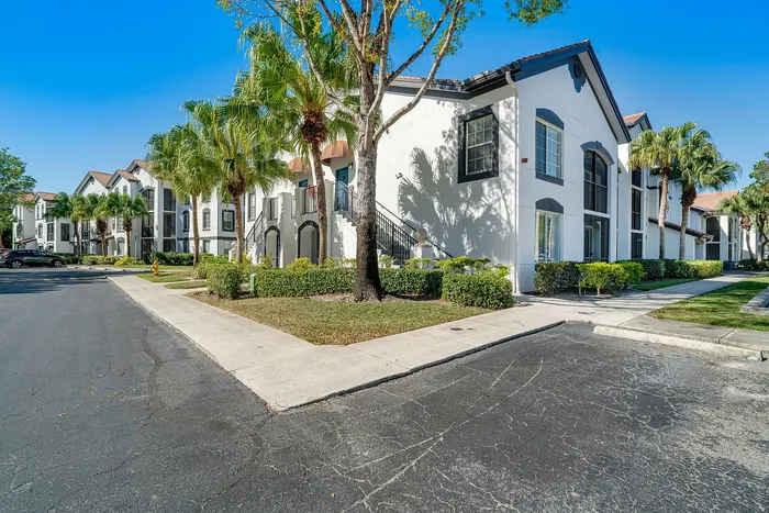 Charming apartment complex with elegant white facades, surrounded by lush palm trees and manicured greenery under a bright blue sky.