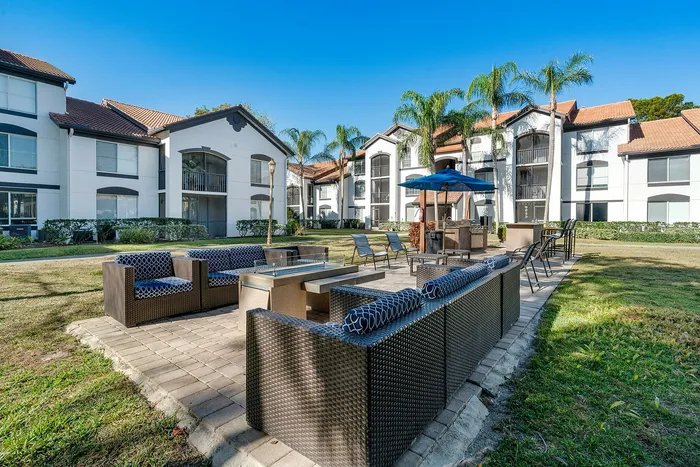 Charming apartment complex with Mediterranean-style architecture, surrounded by lush palm trees and featuring a cozy outdoor seating area with modern furniture and a BBQ grill.