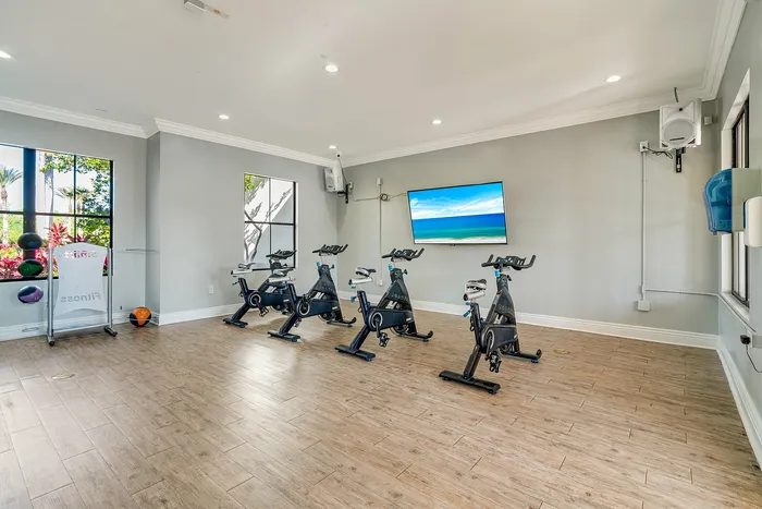 Modern fitness room with four stationary bikes, a sleek wooden floor, vibrant natural lighting, and a wall-mounted TV displaying a scenic view.