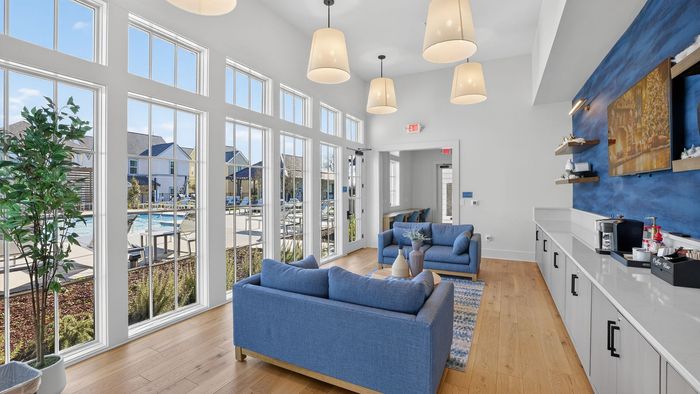 Bright and inviting lounge area featuring cozy blue sofas, decorative pendant lighting, large windows overlooking a sparkling pool, and a modern refreshment bar.