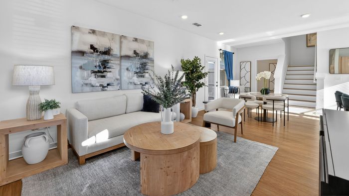 Chic living room featuring a cozy beige sofa, stylish abstract artwork, and vibrant greenery, leading to a sunlit dining area with elegant decor and modern stairs.