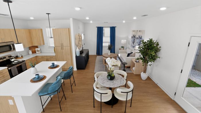 Bright and modern open-concept living space featuring a sleek kitchen with wooden cabinetry, a cozy dining area with a marble round table, and a stylish living room with vibrant decor and plant accents.