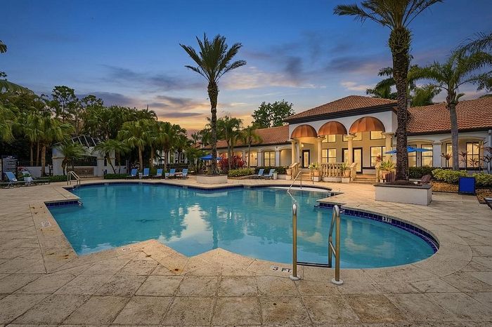 Luxurious resort-style pool area with elegant palm trees, lush landscaping, and a modern clubhouse at sunset.