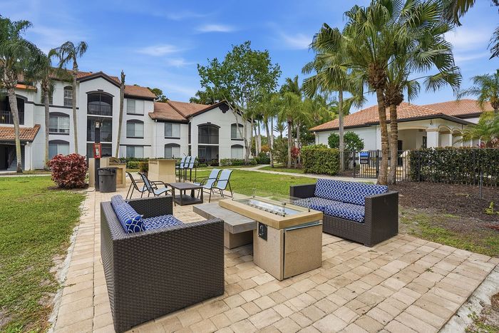 Charming outdoor seating area with stylish wicker furniture, surrounded by lush greenery and palm trees, perfect for relaxation under the sun at a modern apartment complex.