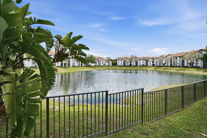 Serene lakeside view of elegant townhomes surrounded by lush greenery under a bright blue sky.