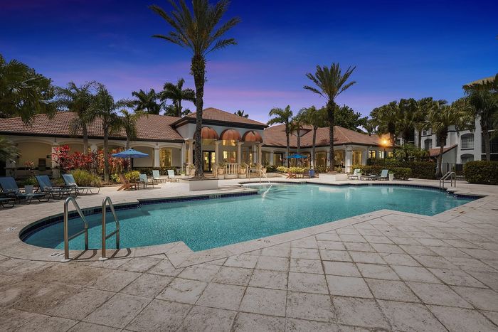Luxurious swimming pool surrounded by palm trees and lounge chairs, set against a beautifully lit clubhouse at twilight.