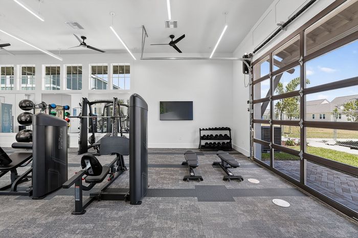 Modern fitness center with state-of-the-art equipment, abundant natural light, and large glass garage door opening to a landscaped outdoor area.