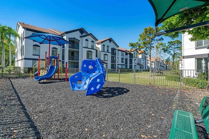 Family-friendly apartment complex featuring modern playground equipment and lush green surroundings under a bright blue sky.