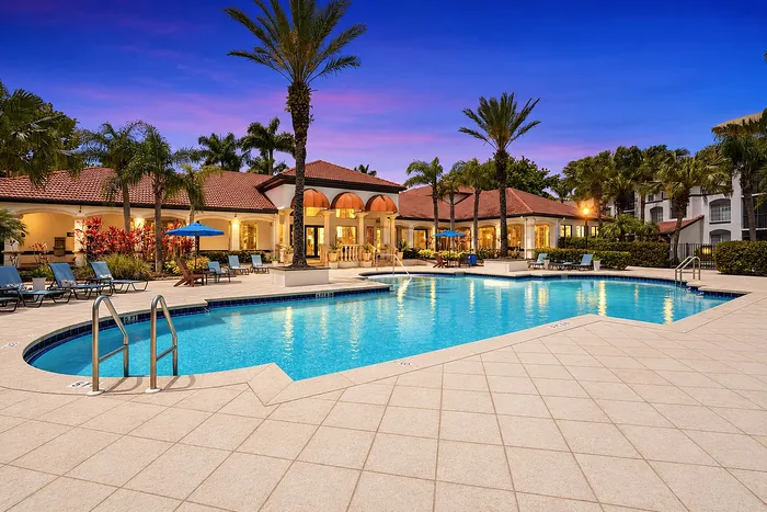 Luxurious resort-style pool surrounded by palm trees and elegant lounge chairs, with a stunning clubhouse in the background at sunset.