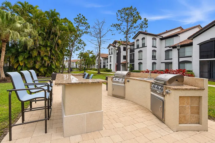 Luxurious outdoor grilling area with modern amenities, surrounded by lush greenery and elegant apartment buildings.