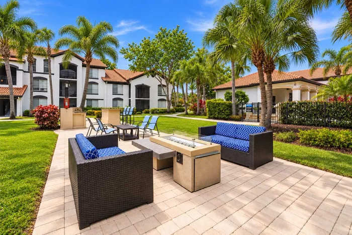 Luxurious apartment complex with stylish outdoor seating, lush green landscaping, and tall palm trees under a bright blue sky.