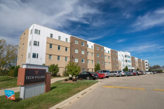 Outdoor sign in foreground with building in background | 	Tech Village | Student Apartments Appleton