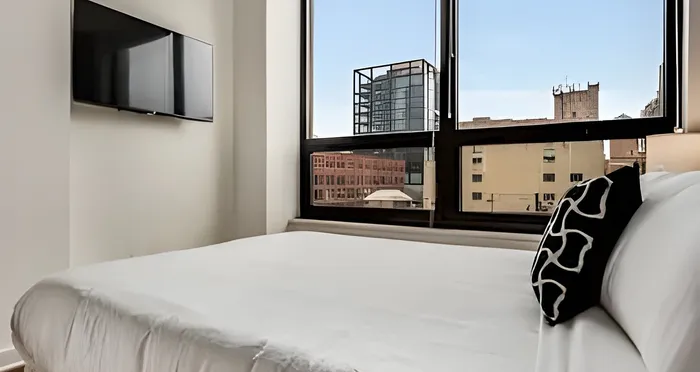 Bright and modern bedroom with a large window offering stunning city views, featuring a sleek mounted TV and comfortable bedding.