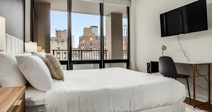 Bright and modern bedroom with a luxurious bed, large windows showcasing a city view, and a sleek wall-mounted TV.