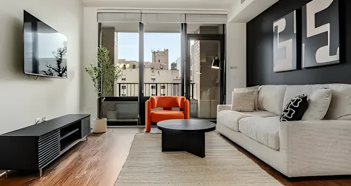 Chic urban living room with a stylish modern design, featuring a cozy white sofa, bold orange chair, and wall-mounted TV against a cityscape backdrop.