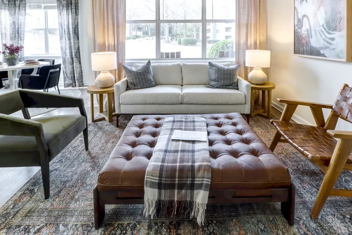 Inviting living room with modern furniture including a plush sofa, stylish armchairs, and a large tufted ottoman, accented by warm lighting and large windows.