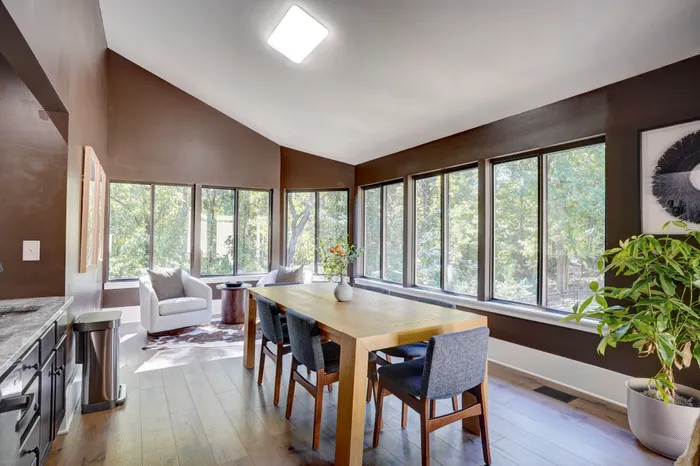 Bright dining area with a large wooden table and chairs, surrounded by expansive windows offering lush garden views.