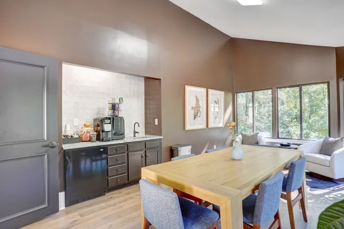 Inviting dining area with a modern kitchenette, elegant wooden table, and large windows offering lush green views, perfect for entertaining.